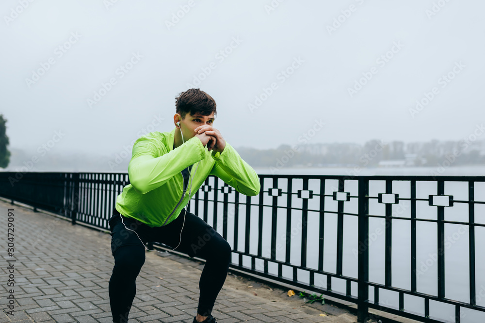Outdoor image of young athletic man exercising squats at city lake background. Fit male doing squats outdoors in morning in the park on fog background. Sport, lifestyle and people concept. Outdoor image of young athletic man exercising squats at city lake background. Fit male doing squats outdoors in morning in the park on fog background. Sport, lifestyle and people concept.