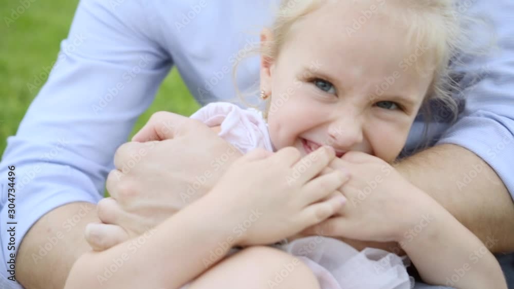 dad hugs his little blonde adorable daughter in pink dress sitting on the meadow, close up dad hugs his little blonde adorable daughter in pink dress sitting on the meadow, close up