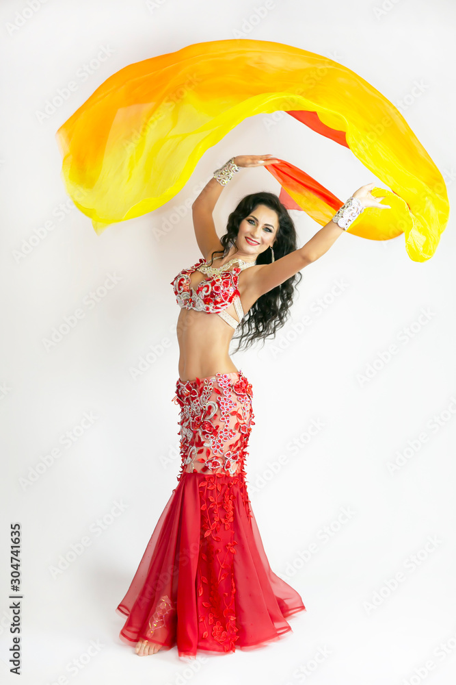 The dancer of east dances with bright scarfs fabrics. A brunette woman performs a belly dance waving in air with colored shawls on a white background. The dancer of east dances with bright scarfs fabrics. A brunette woman performs a belly dance waving in air with colored shawls on a white background.