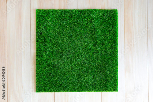 Grass square on wooden background. A piece of green grass in the shape of a square is laid out on a light wooden background.