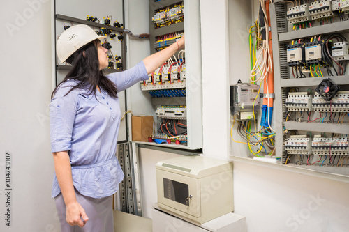 Female engineer in white helmet switches on circuit breakers in electrical box. Professional girl checks the switchboard of the enterprise. The specialist works in the electrical distribution panel.