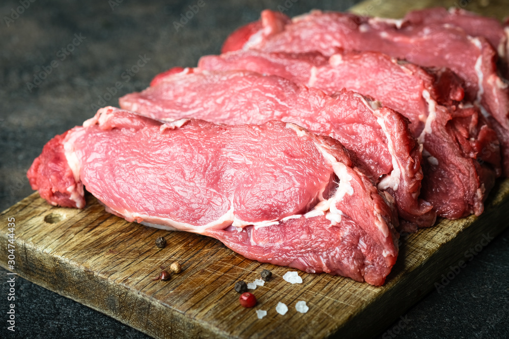 Fresh raw beef, cut into steaks on a wooden board with vegetables, herbs and spices on a wooden board on a dark background. Fresh raw beef, cut into steaks on a wooden board with vegetables, herbs and spices on a wooden board on a dark background.