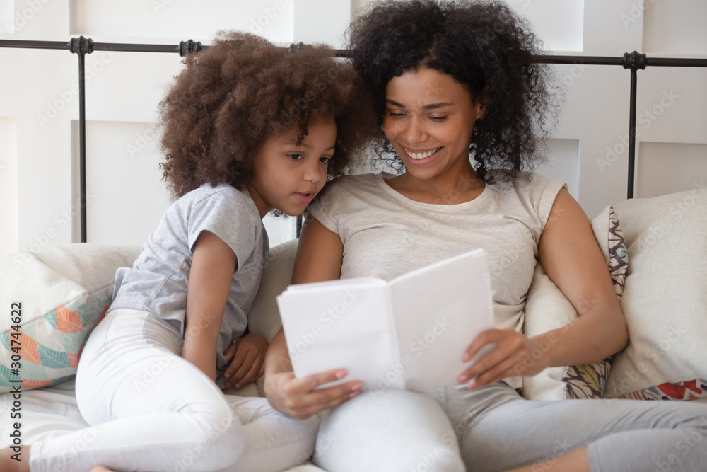 Happy biracial mom and daughter reading in bedroom together