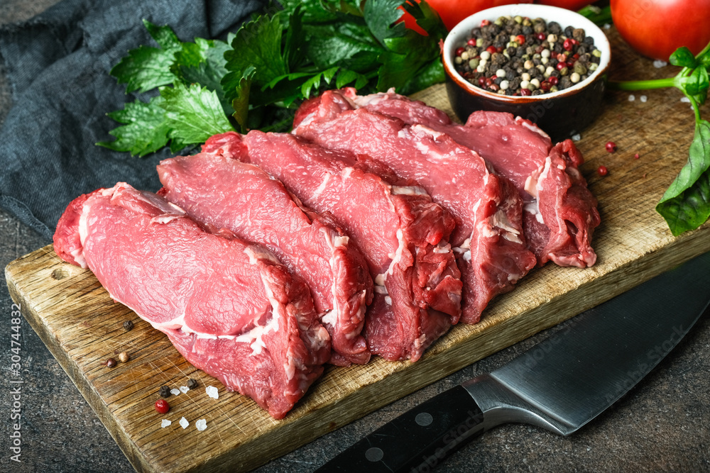 Fresh raw beef, cut into steaks on a wooden board with vegetables, herbs and spices on a wooden board on a dark background.