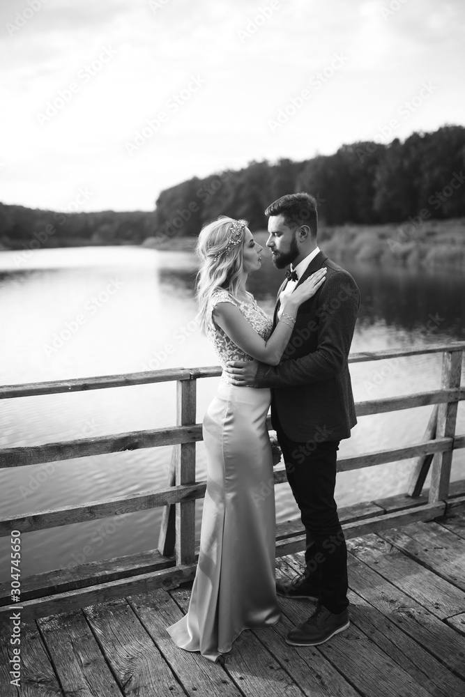 Black and white foto stylish couple of newlyweds posing on a bridge on wedding day. Handsome bearded groom admires and kisses pretty bride. Together. The concept of youth, love, fashion and lifestyle.