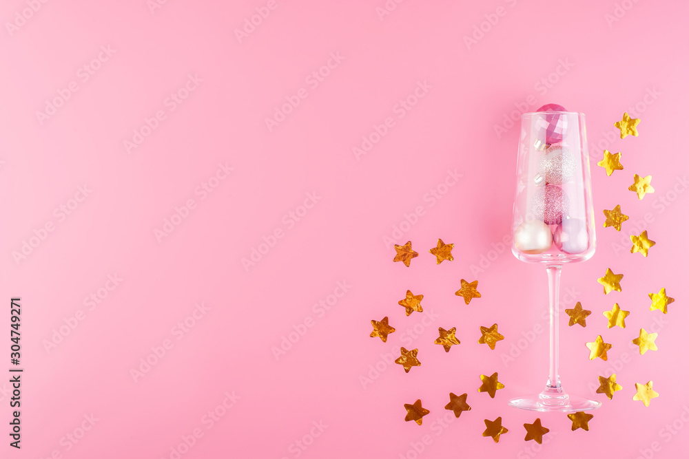 A glass of champagne is filled with pink and gold shiny decorations in the form of balls on a pink pastel background A glass of champagne is filled with pink and gold shiny decorations in the form of balls on a pink pastel background