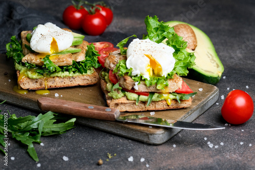 Delicious sandwich with avocado and poached egg, cherry tomatos and arugula on a dark background.