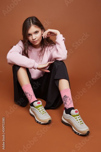 Full length shot of a dark-haired girl, sitting on a floor. She wearing pink sweatshirt, black culottes, blue artwork socks with Queen Victoria print and white sneakers. Her legs is benting.   photo