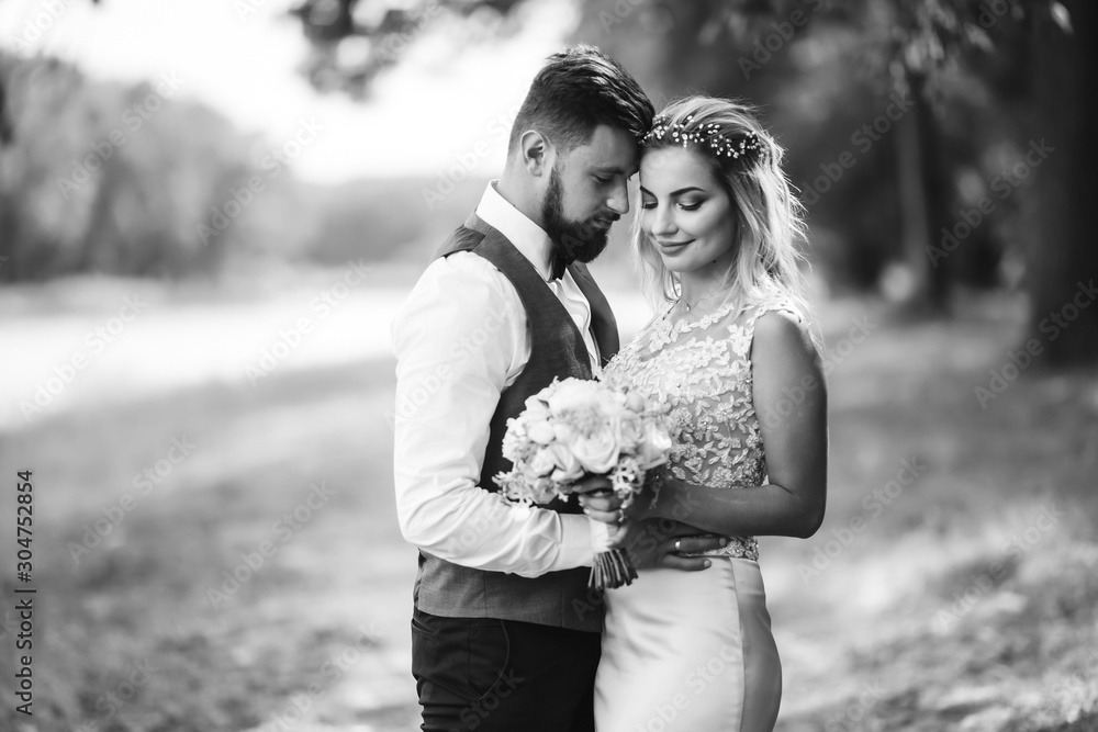 Fototapeta premium Sensual Black and white foto of bride and groom. Stylish couple of happy newlyweds posing and kisses in the park on their wedding day. Together. The concept of youth, love, fashion and lifestyle. 