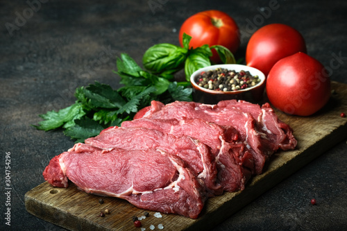 Fresh raw beef, cut into steaks with vegetables, herbs and spices on a wooden board on a dark background.