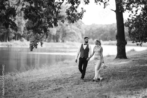 Sensual Black and white foto of bride and groom. Stylish couple of happy newlyweds posing in the park on their wedding day. Handsome bearded groom admires and kisses pretty bride. Together.