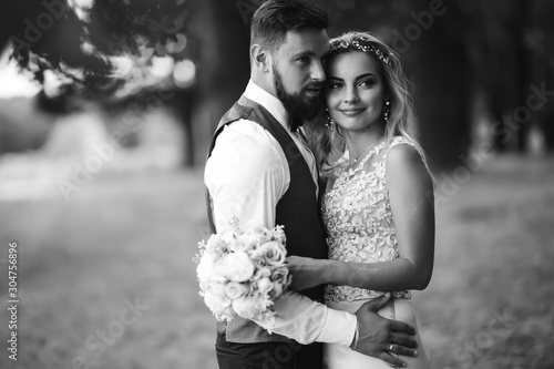 Sensual Black and white foto of bride and groom. Stylish couple of happy newlyweds posing in the park on their wedding day. Handsome bearded groom admires and kisses pretty bride. Together.