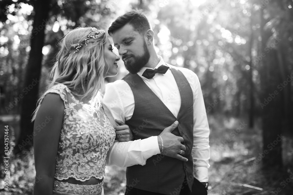 Fototapeta premium Sensual Black and white foto of bride and groom. Stylish couple of happy newlyweds posing in the park on their wedding day. Handsome bearded groom admires and kisses pretty bride. Together.