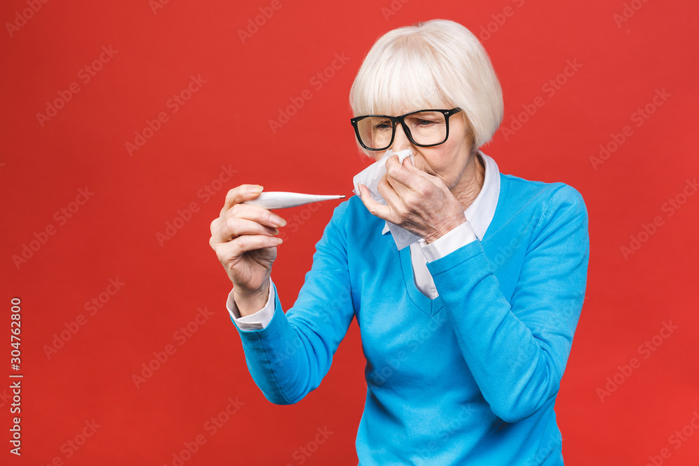 Ill allergic old mature woman blowing runny nose got hay fever rhinitis allergy flu, sick middle aged senior lady sneezing in tissue holding handkerchief isolated on red studio background.