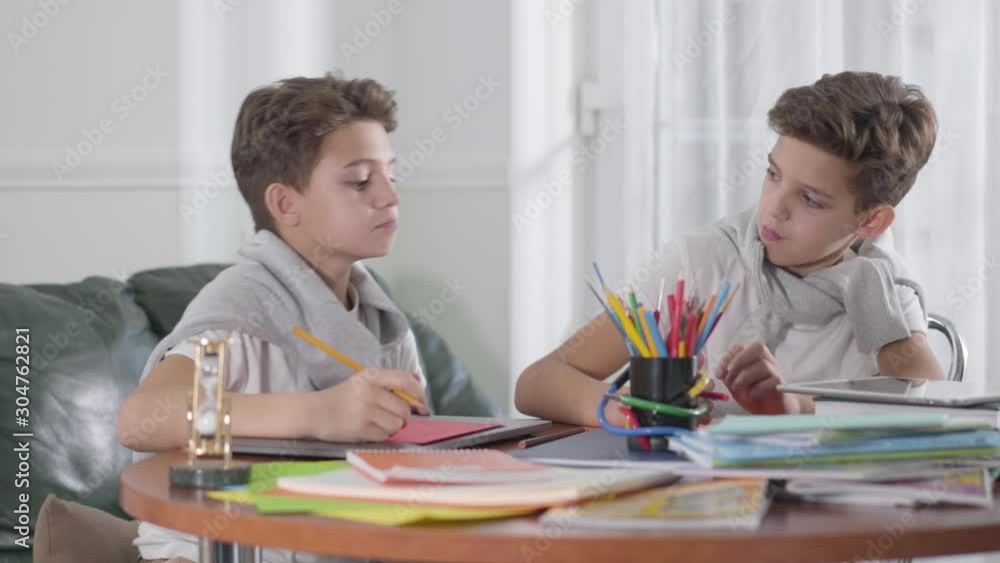 Two Caucasian twin brothers sitting at the table and studying. One boy trying to cheat, his sibling closing his exercise book. Schoolchildren doing homework at home. Education concept. Two Caucasian twin brothers sitting at the table and studying. One boy trying to cheat, his sibling closing his exercise book. Schoolchildren doing homework at home. Education concept.