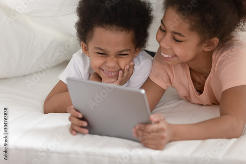 Cheerful sister and brother lying on bed using tablet computer