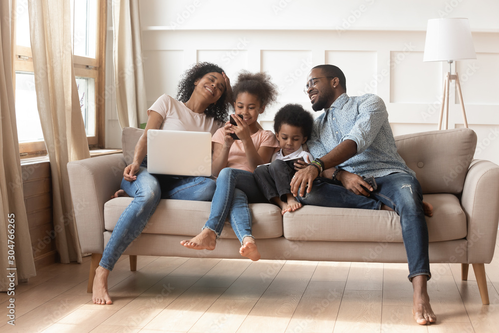 Cheerful couple and kids with gadgets sitting on couch