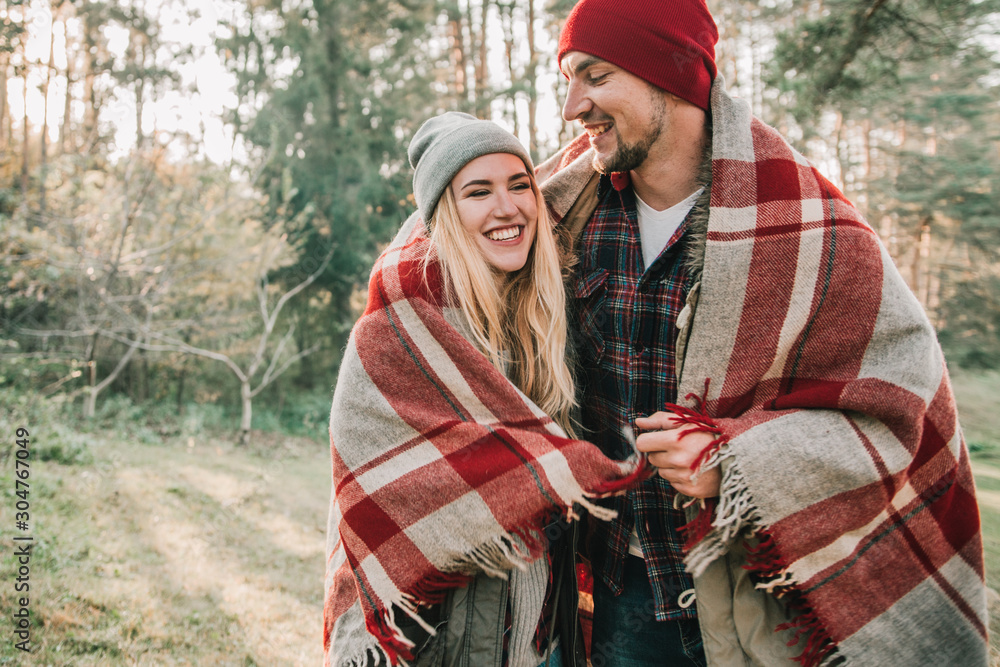 Couple huggings travelers covered with plaid in the forest. Concept of trekking, adventure and seasonal vacation.