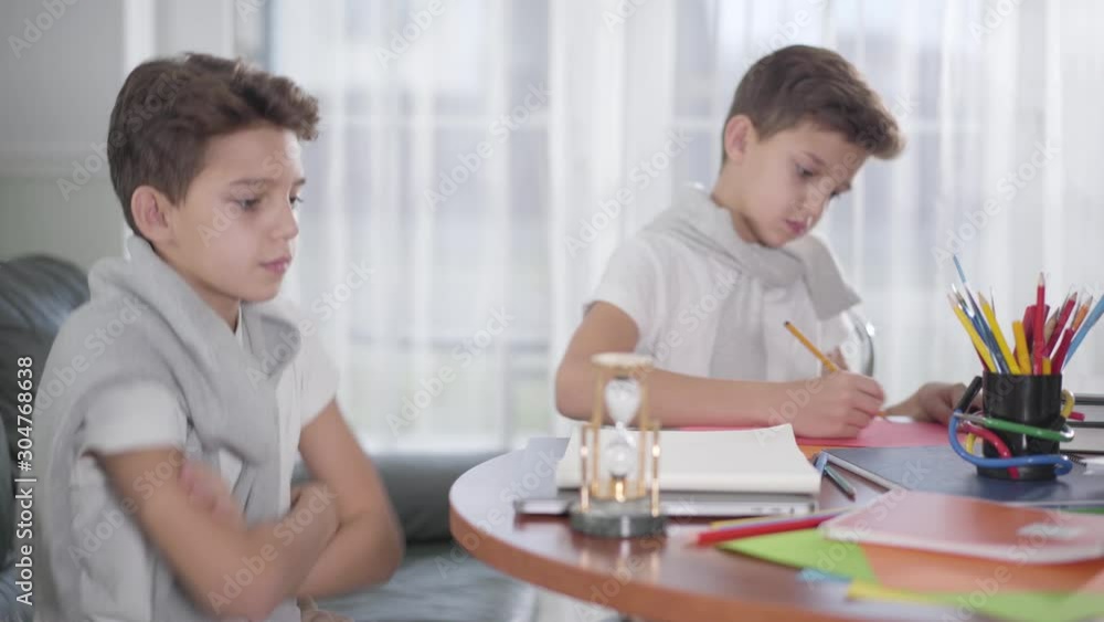 Side view of little Caucasian boy waiting for twin brother to drawing the picture and giving high five. Smart siblings doing homework together at home. Generation Z, bright mind, education concept. Side view of little Caucasian boy waiting for twin brother to drawing the picture and giving high five. Smart siblings doing homework together at home. Generation Z, bright mind, education concept.