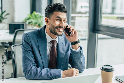 cheerful businessman talking on smartphone while sitting at workplace in office