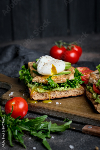 Delicious sandwich with avocado and poached egg, with green leaves and tomatoes on a dark background. Healthy breakfast.