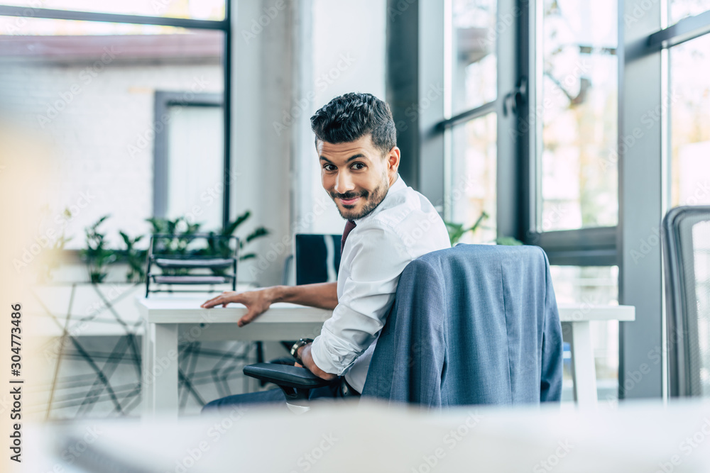 selective focus of cheerful businessman smiling at camera while sitting at workplace