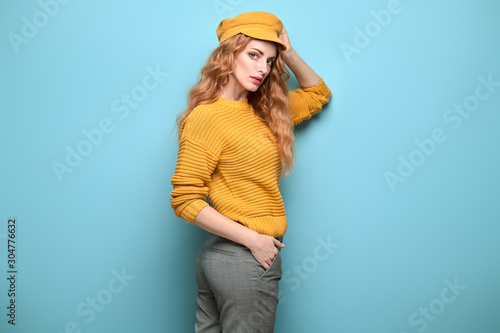 Fashionable woman in Trendy orange outfit, stylish hairstyle, makeup. Young redhead in jumper, yellow cap. Sensual beautiful model girl in stylish pants, fashion beauty concept on blue