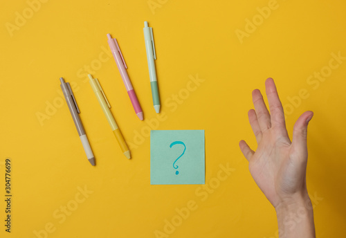 Surprised female hand, memo piece of paper with a question mark and pens on yellow background