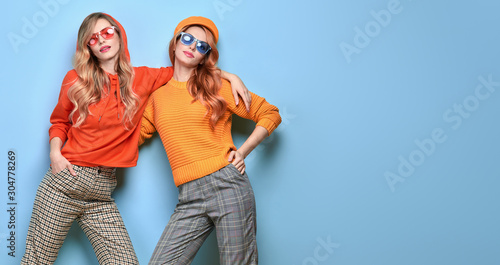 Two Lovable embracing fashionable woman sisters in Trendy orange yellow outfit. Studio shot of Carefree beautiful funny stylish friends smiling on blue. Happy fashion girl, colorful positive banner