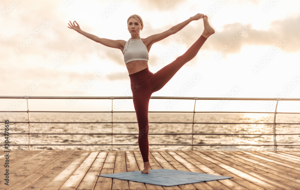 Obraz premium Healthy woman practising yoga on seaside promenade. Sport woman standing in yoga pose with one leg raised at sunrise. Utthita Hasta Padangusthasana