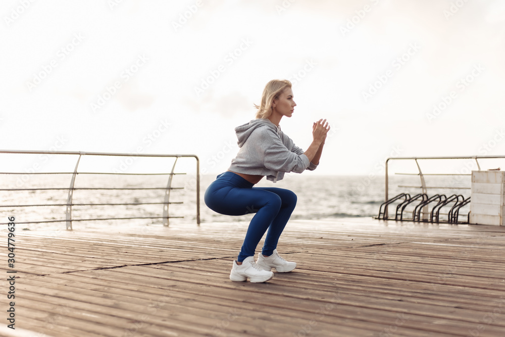 Healthy woman training on seaside promenade. Sportswoman in sportwear doing squat exercise on the beach at sunrise