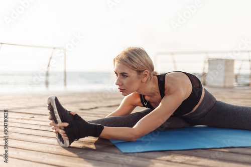 Healthy woman training on seaside promenade. Fitness woman in sportswear doing leg stretching while sitting on a mat on the beach at sunrise