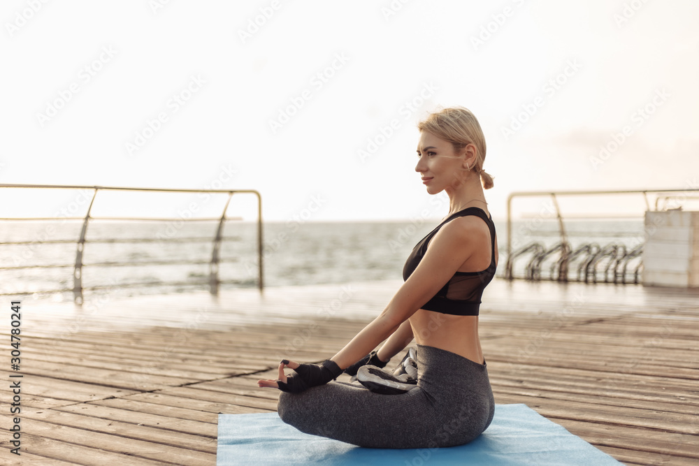 Practice yoga in the morning outdoors. Young woman yogi in sportswear is sitting in lotus position on the beach at sunrise. Inner harmony