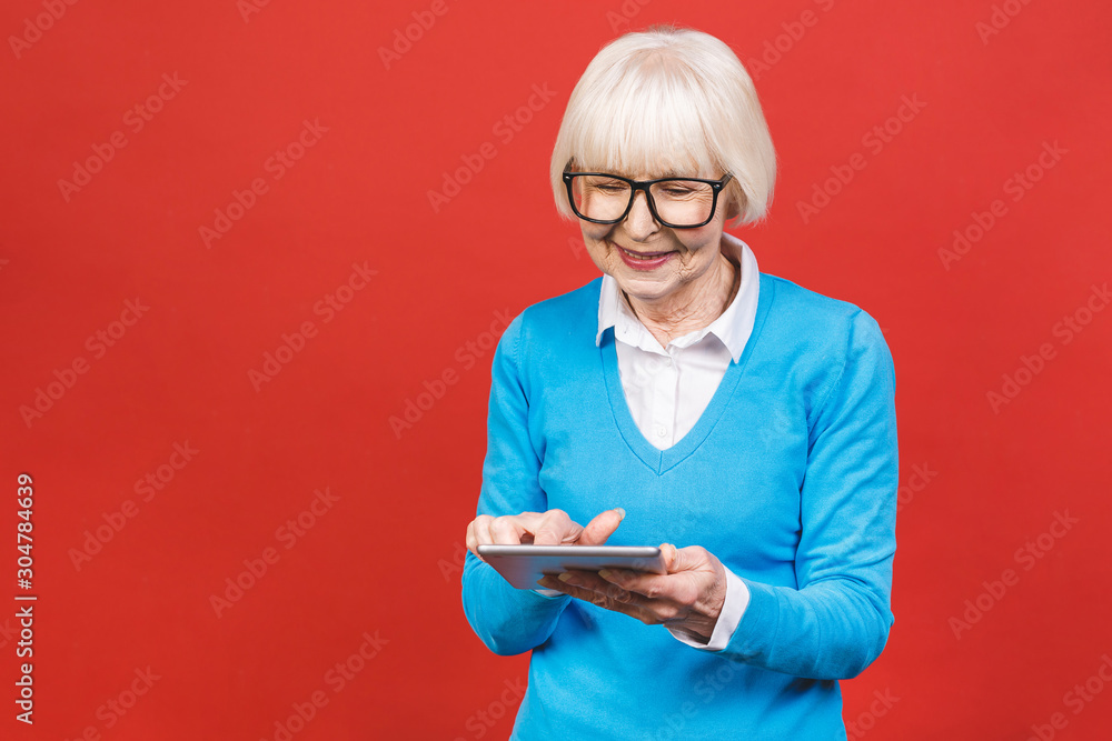 Grey haired old business woman reading browsing using holding tablet. Isolated over red background. Grey haired old business woman reading browsing using holding tablet. Isolated over red background.
