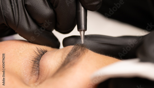 Young attractive man eye close-up with the master hand in black gloves and permanent makeup machine with a needle.