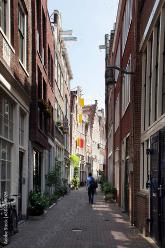 View of man walking on a narrow street in Amsterdam. Historical, traditional and typical buildings and many plants are in the view. It is a sunny summer day.