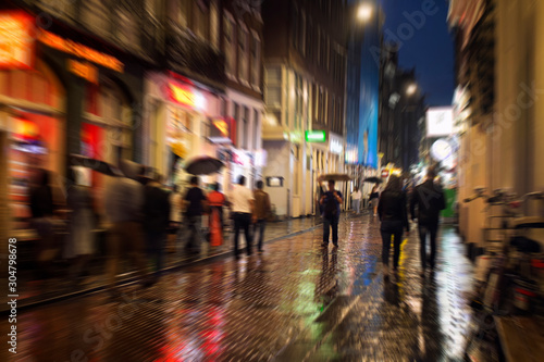 Blurry motion image of people walking on Warmoesstraat street in Amsterdam. It is one of the main shopping streets with cafes, restaurants and shops. It is a rainy summer night. Youth culture concept © theendup
