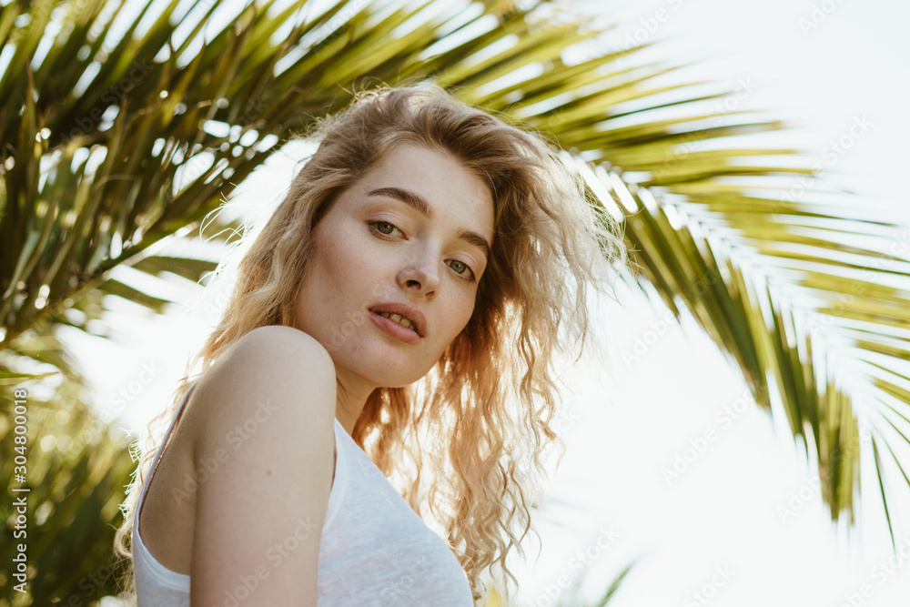 girl inquiringly looks at the camera from top to bottom, standing under a palm branch girl inquiringly looks at the camera from top to bottom, standing under a palm branch