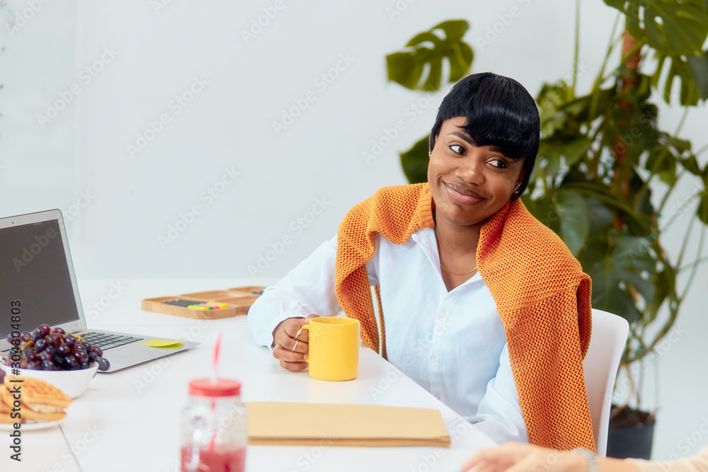 Charming african lady wearing casual clothes sit with cup of tea in modern office, break time during work. Creative business people Charming african lady wearing casual clothes sit with cup of tea in modern office, break time during work. Creative business people