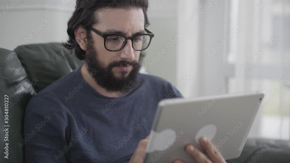 Portrait of concentrated Caucasian man in eyeglasses using tablet. Young bearded businessman dealing with issues on weekends at home. Distant work, freelance, overworking. Portrait of concentrated Caucasian man in eyeglasses using tablet. Young bearded businessman dealing with issues on weekends at home. Distant work, freelance, overworking.