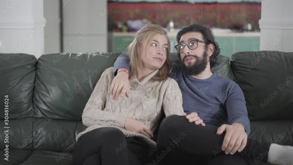 Happy Caucasian couple sitting on couch at home and discussing renovation works. Young bearded man in eyeglasses embracing beautiful woman and talking with her. Happy family planning future. Happy Caucasian couple sitting on couch at home and discussing renovation works. Young bearded man in eyeglasses embracing beautiful woman and talking with her. Happy family planning future.