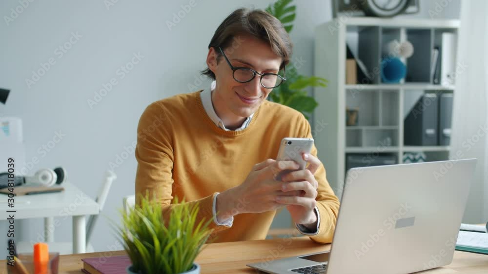 Good-looking young man using smartphone in office at desk smiling having fun enjoying communication online. Modern technoology and youth concept. Good-looking young man using smartphone in office at desk smiling having fun enjoying communication online. Modern technoology and youth concept.