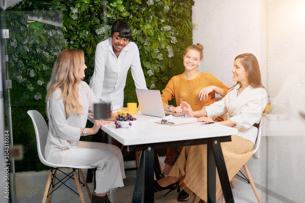 Confident young african lady in white shirt stand talking to caucasian young women, teaching business mentoring, discuss strategy in modern office Confident young african lady in white shirt stand talking to caucasian young women, teaching business mentoring, discuss strategy in modern office