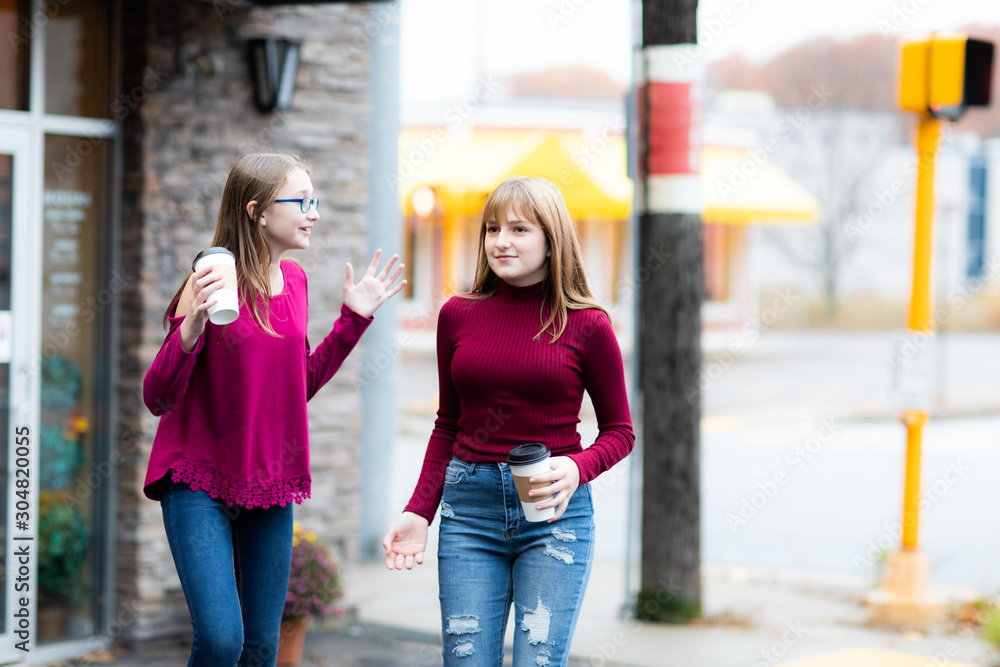 Two young teenager girls going from coffee store. Sisters talking and arguing. Holding coffee cups and drinking.  Casual style, city street