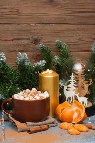 Cup of cocoa with marshmallow, cinnamon rolls, tangerine, burning candles and christmas tree garland near wooden wall. 