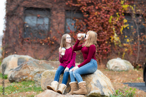 Two young teenager girls sitting on the rock, talking, argue,drinking coffee. Sisters, friends happy smiling, laughing, talking. Fall autumn time.