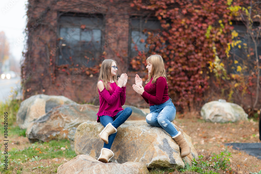 Two young teenager girls sitting on the rock, drinking coffee. Sisters, friends happy smiling, laughing, talking. Fall autumn time.Playing clapping hands Two young teenager girls sitting on the rock, drinking coffee. Sisters, friends happy smiling, laughing, talking. Fall autumn time.Playing clapping hands