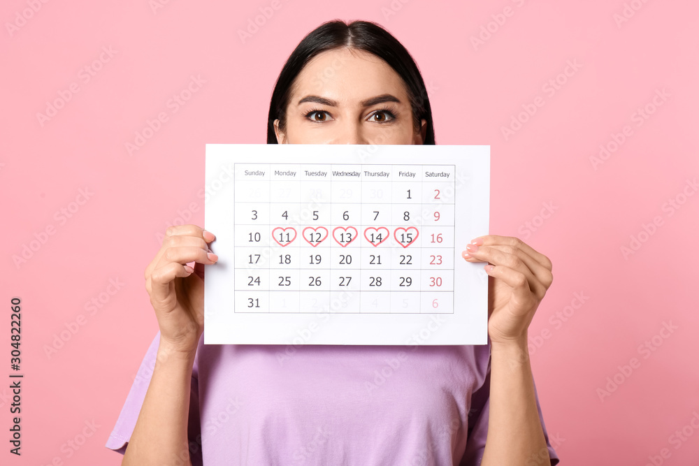 Young woman holding calendar with marked menstrual cycle days on pink background Young woman holding calendar with marked menstrual cycle days on pink background