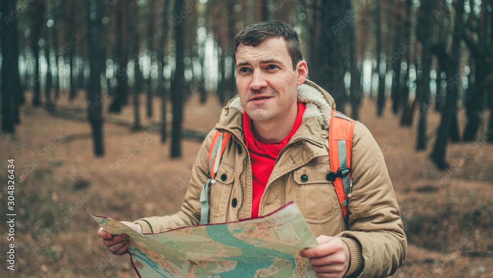 A traveling man with map in woods. A portrait of the handsome man with a backpack, standing near a tree in a cold weather.