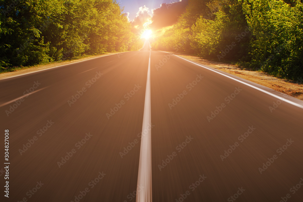 An empty countryside road in motion with green trees against a sky with sunset